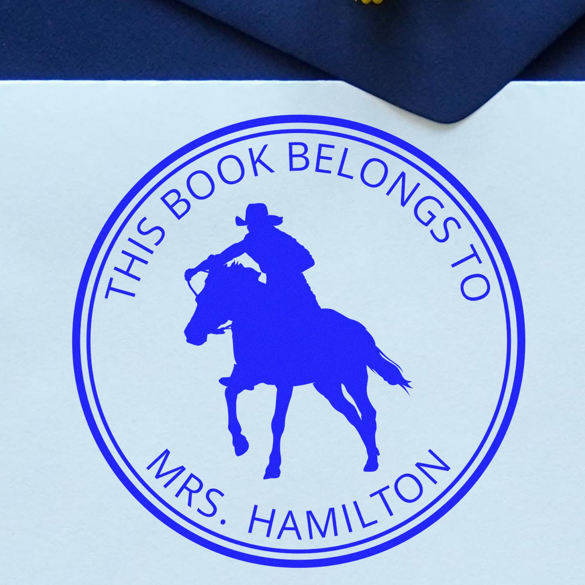 Blue circular book ownership stamp seal with cowboy on horse and text This Book Belongs to Mrs. Hamilton.