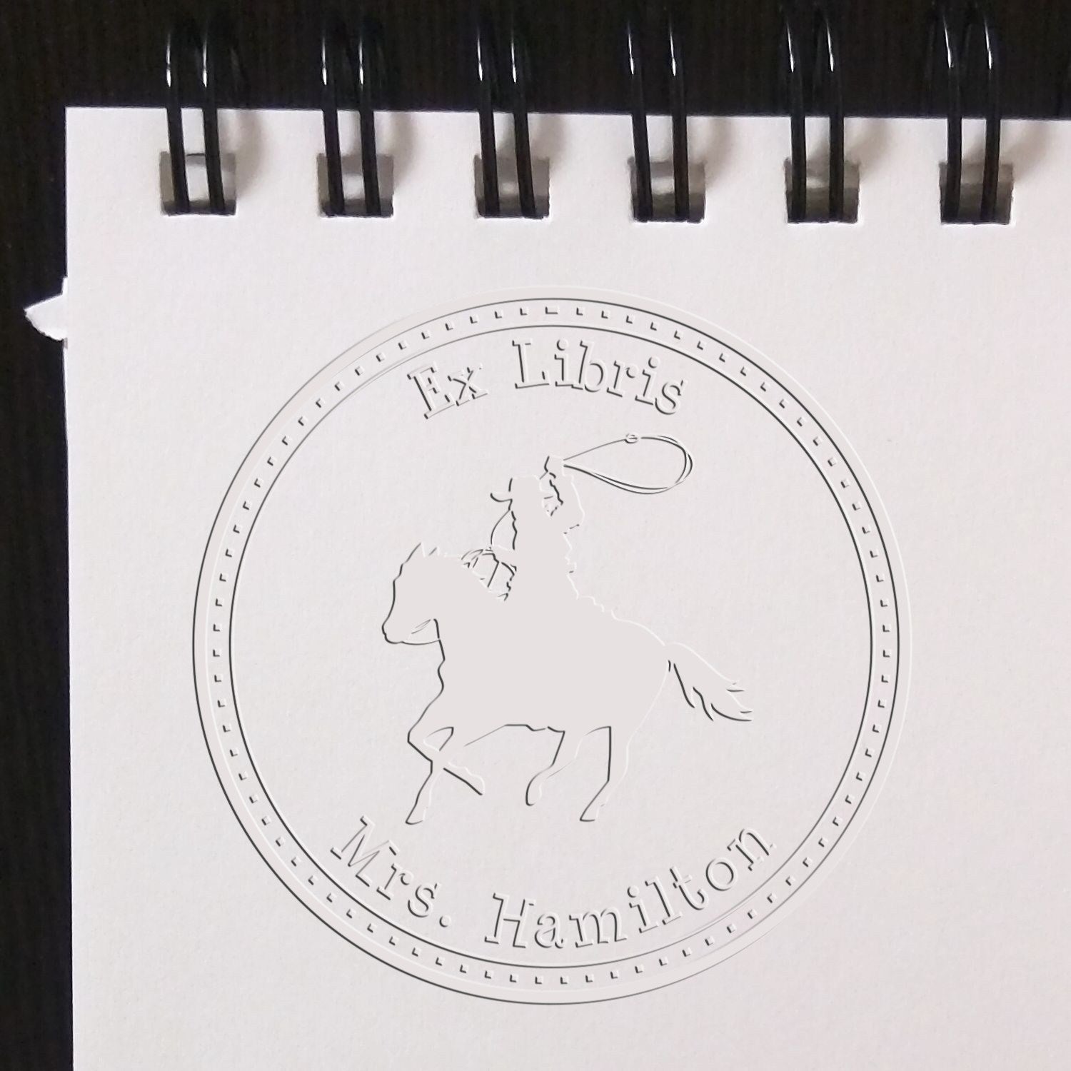 Embossed white circular ex libris stamp seal in a spiral notebook, featuring a horse and rider with Ms. Hamilton text.