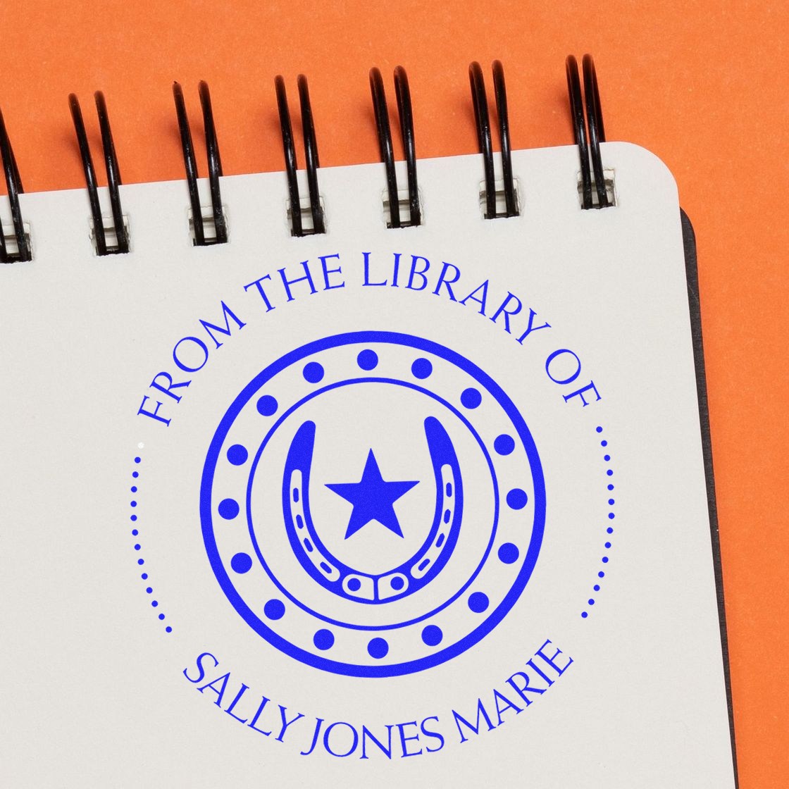 Blue custom library stamp and seal with horseshoe and star on a white spiral notebook over orange background.