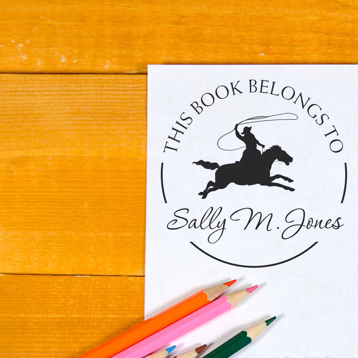 Custom black book-belongs stamp seal on white paper, with cowboy horse silhouette and colorful pencils on orange wood.