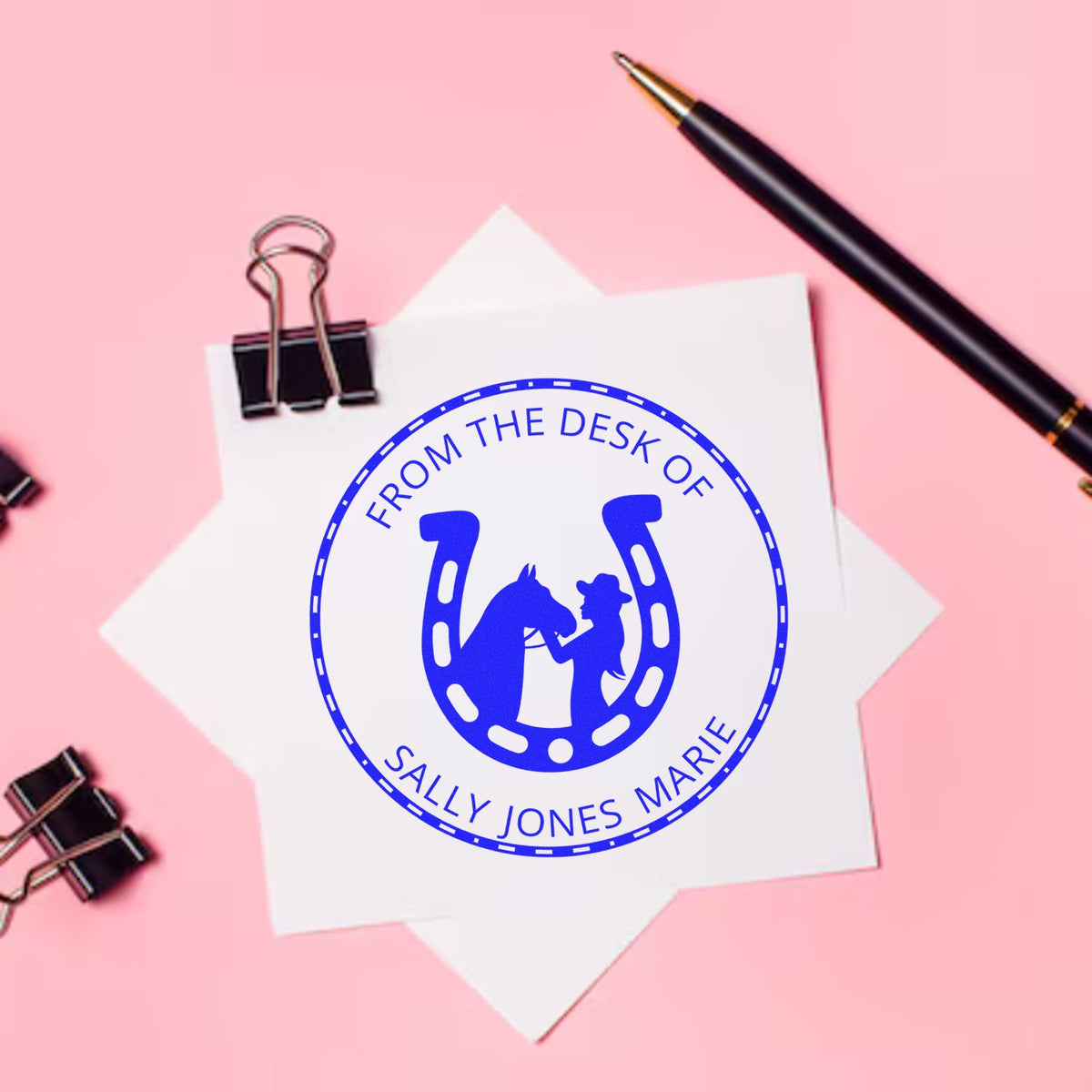 Blue custom horse-and-rider stamp seal on white paper, with black pen and binder clips on a pink background.