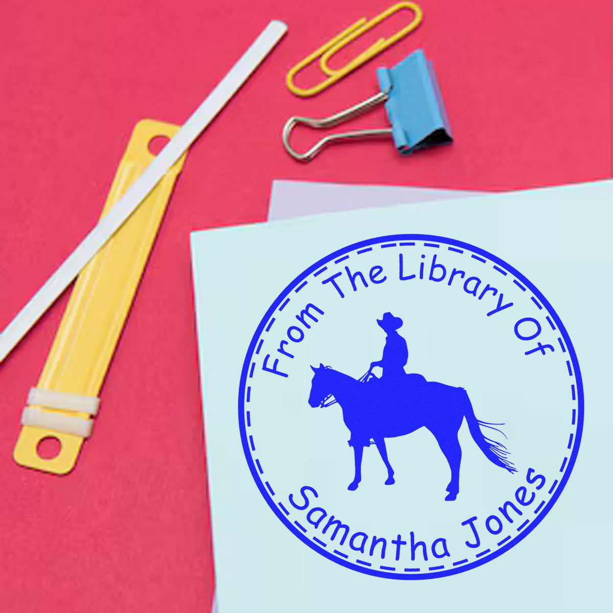 Blue custom library seal on pale blue paper with cowboy on horse, on a red background with paper clips and binder clip.