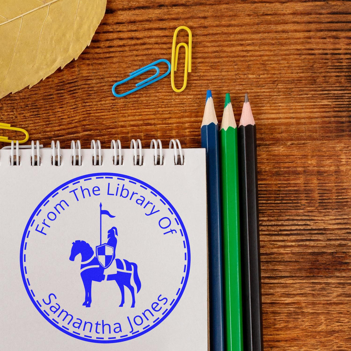 Blue custom library stamp and seal on a spiral notebook, beside colored pencils and paper clips on a wooden desk.