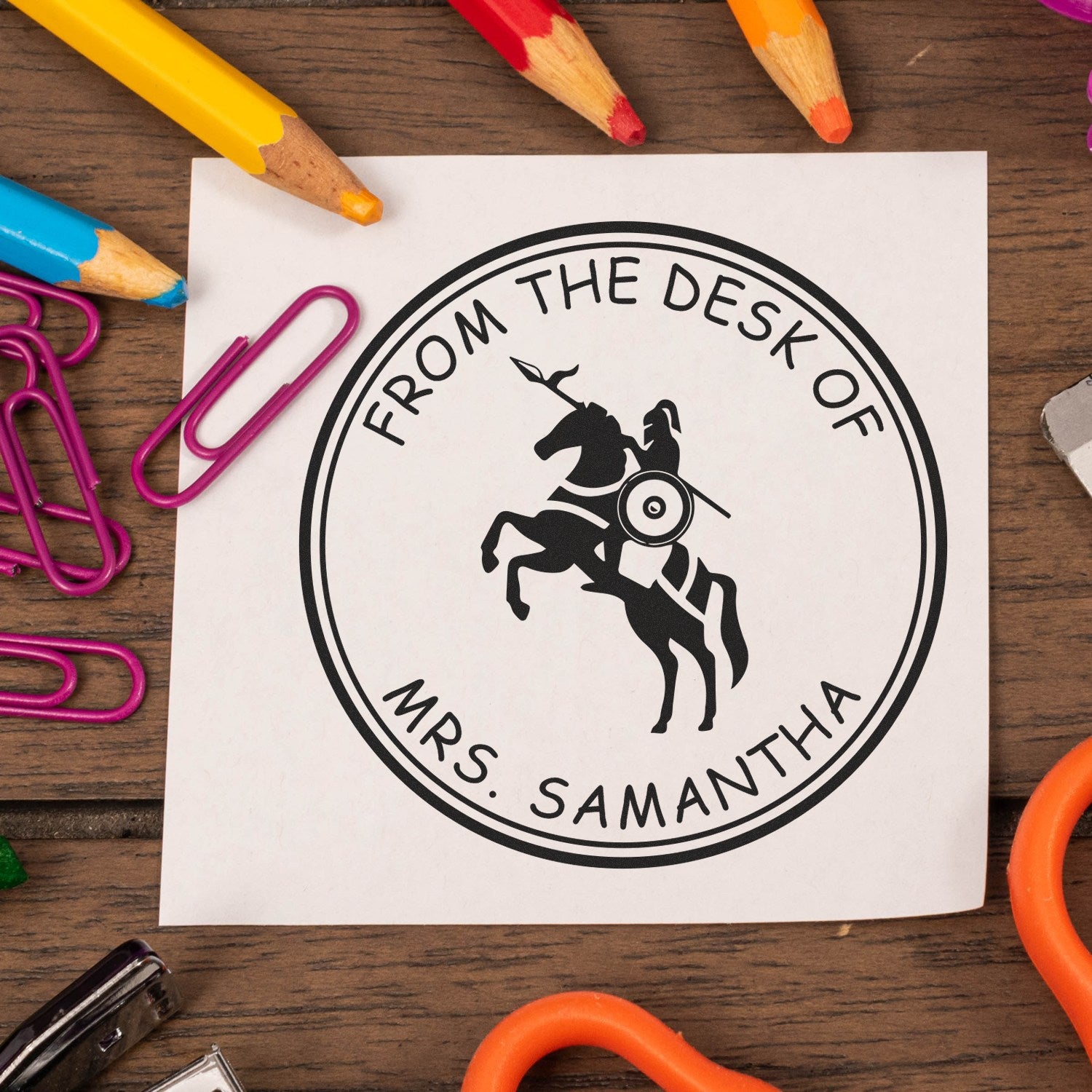 Black custom desk seal stamp reading "From the Desk of Mrs. Samantha" with horse logo, on white paper with colorful pencils and paper clips.