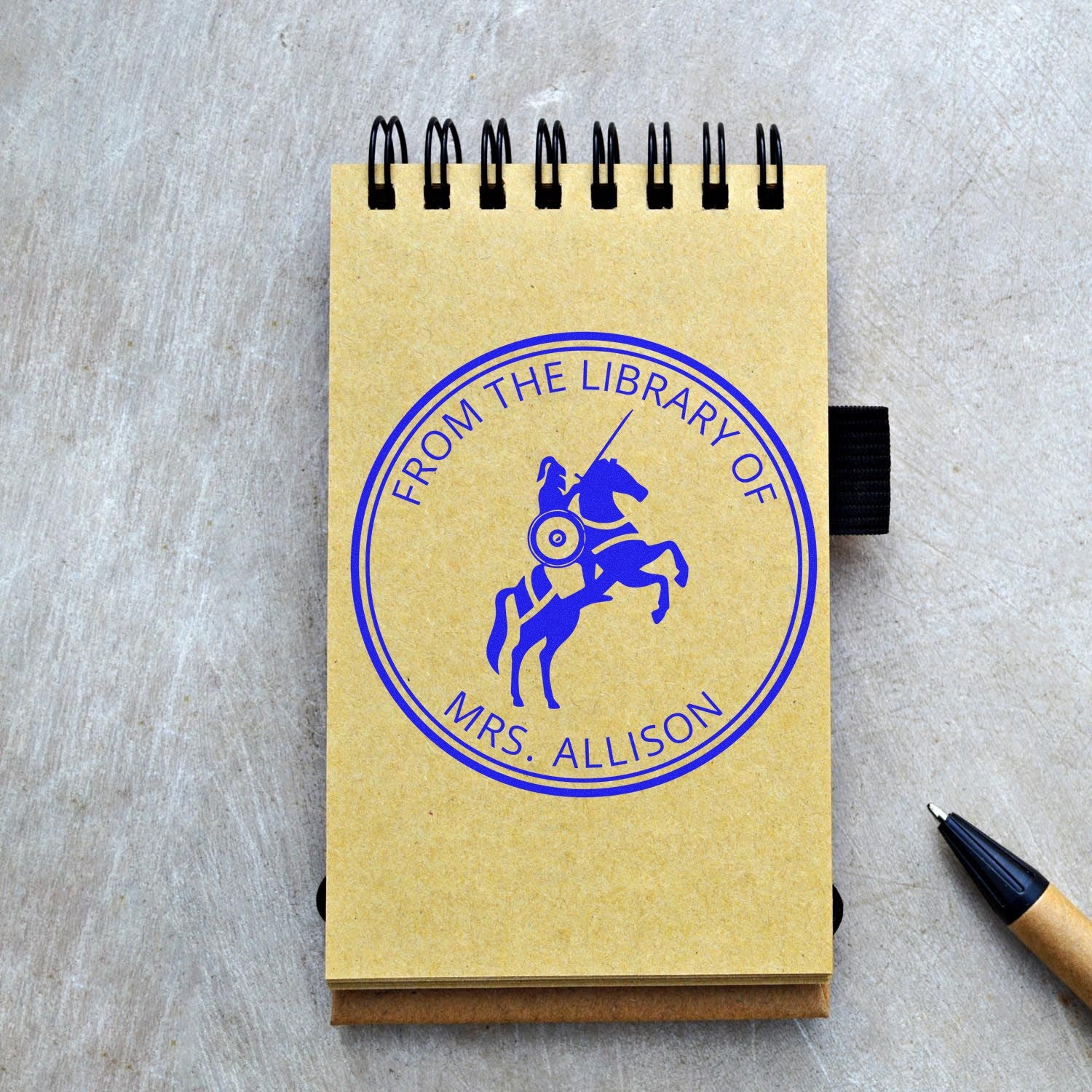 Yellow spiral notebook with blue custom library stamp seal, knight emblem, and black pen on gray wood surface.