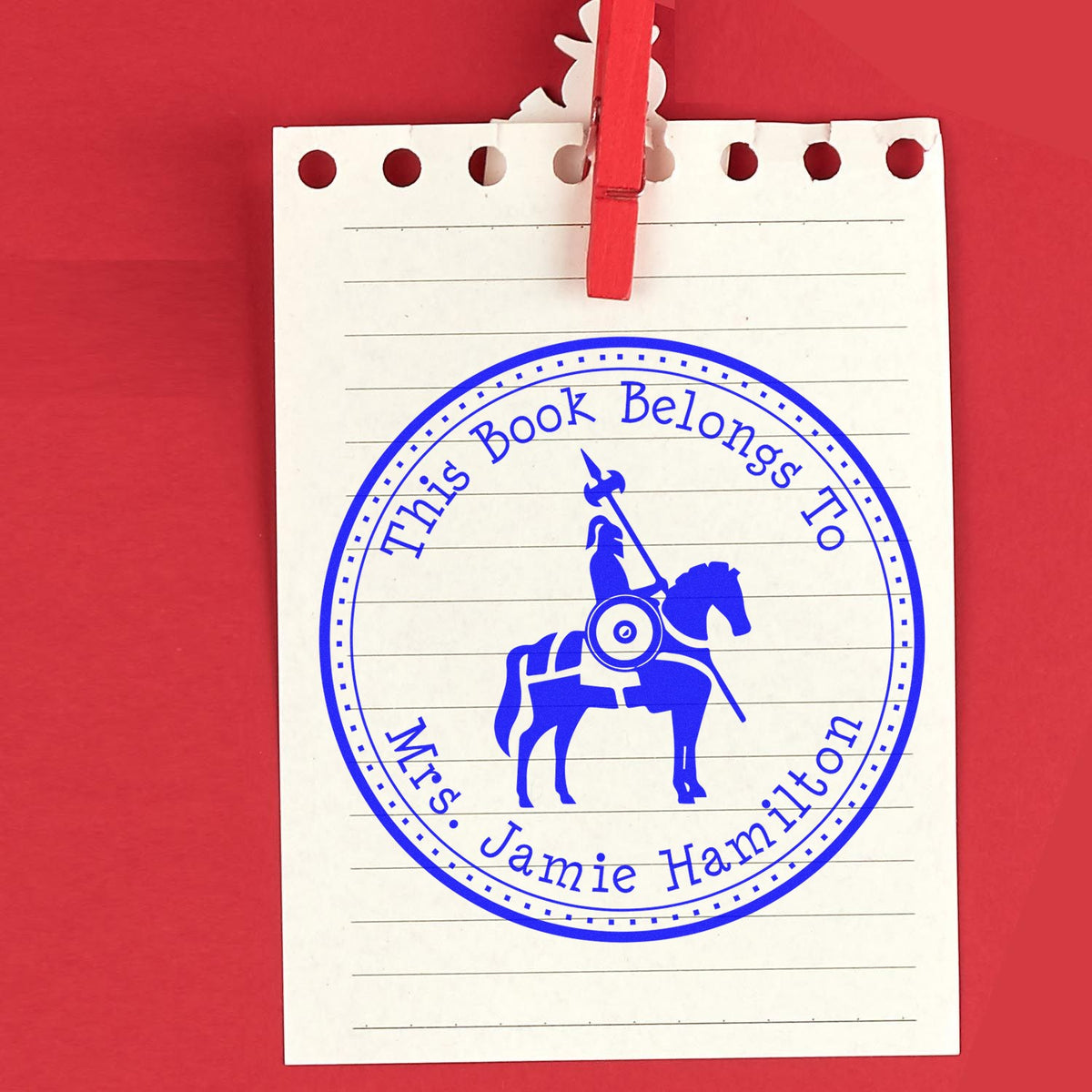 Blue custom book seal stamp reading This Book Belongs To Mrs. Jamie Hamilton on a lined notepad, clipped with red clothespin.