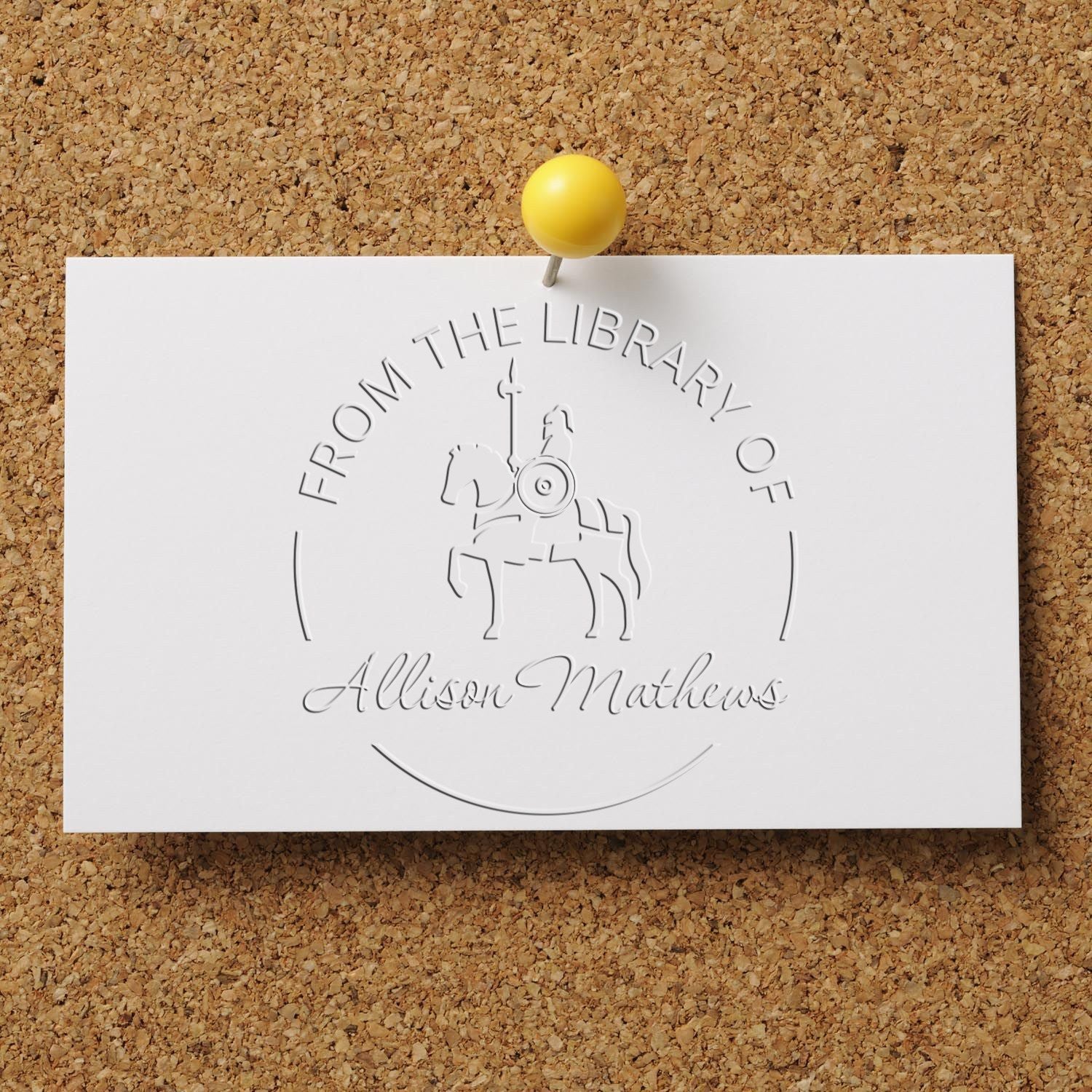 White embossed stamp and seal card with silver library crest and script name, pinned to corkboard with a yellow pushpin.