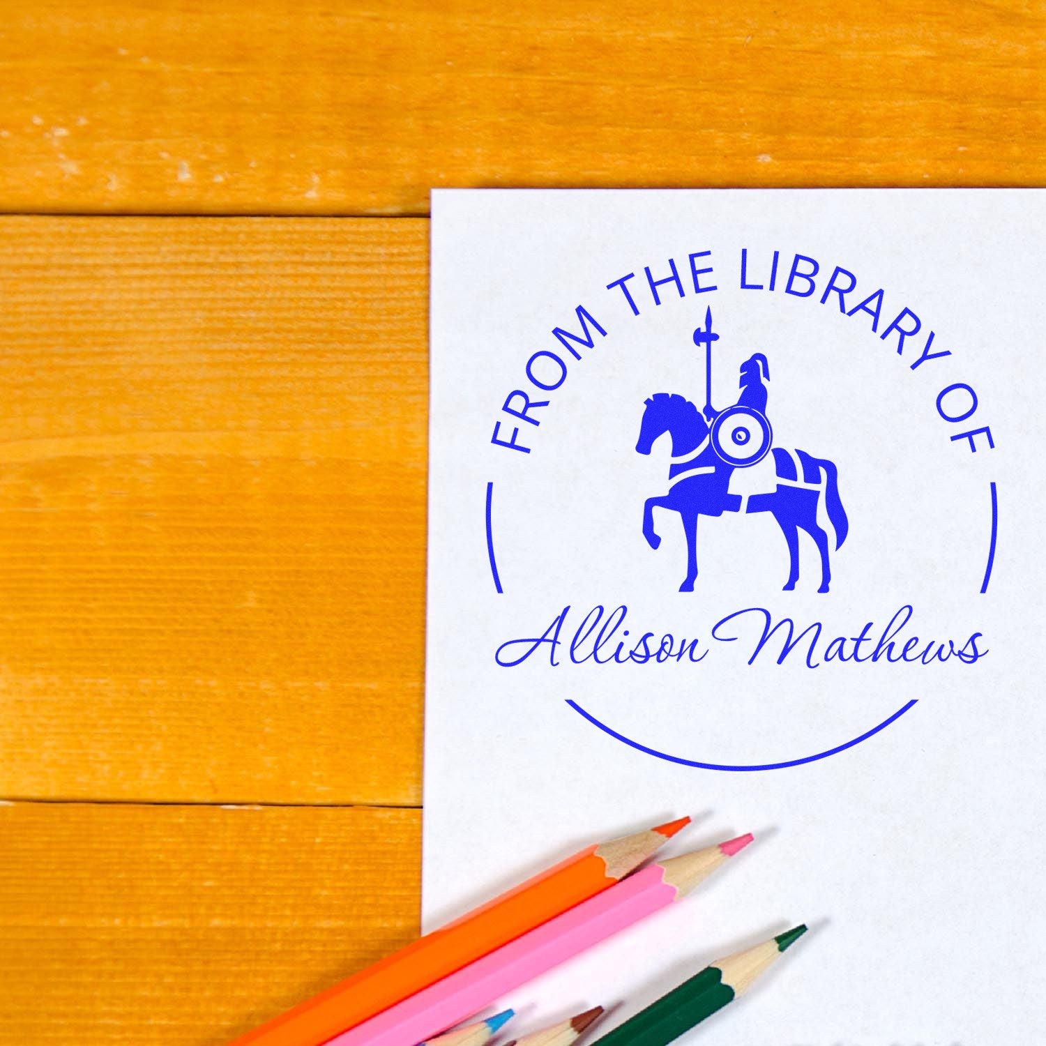 Custom blue library seal stamp on white paper with horse emblem and Allison Matthews name, beside colored pencils.