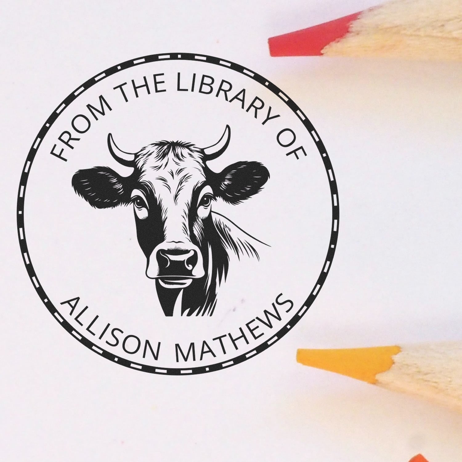 Black circular library stamp seal with cow head and From the Library of Allison Mathews, on white with red and yellow pencils.
