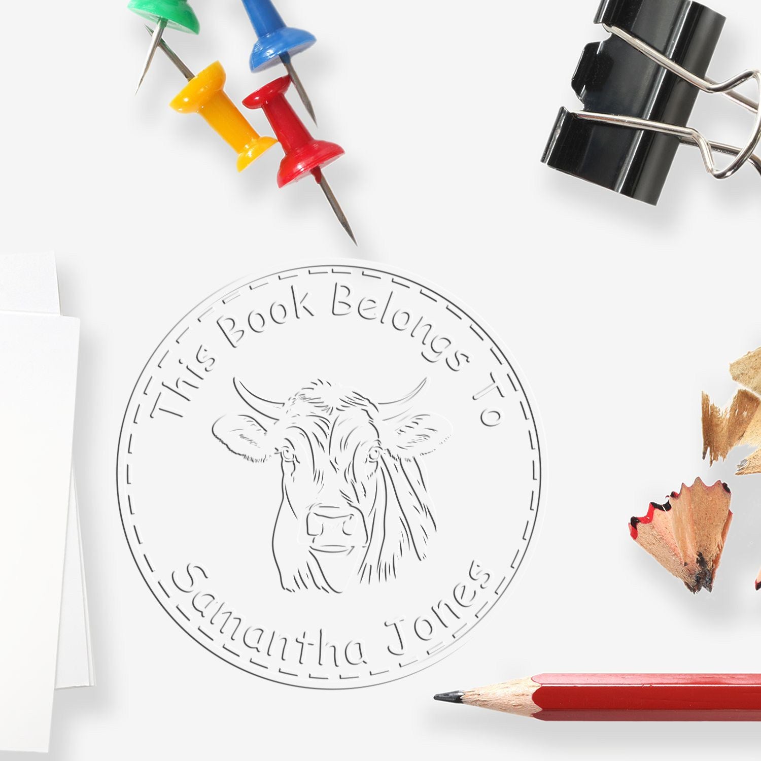 White embossing seal with cow and This Book Belongs to Samantha Jones, surrounded by colorful push pins, binder clip, and pencils