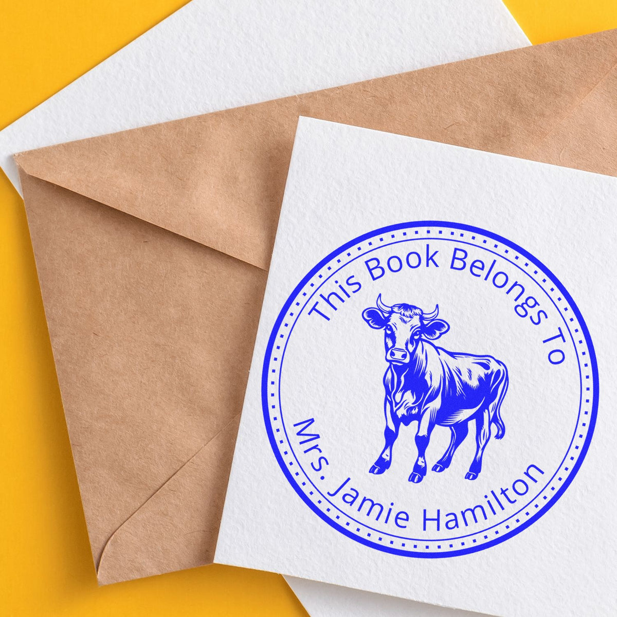 Blue book ownership seal stamp on white card, beside brown envelope on yellow background, with cow and Mrs. Jamie Hamilton text.