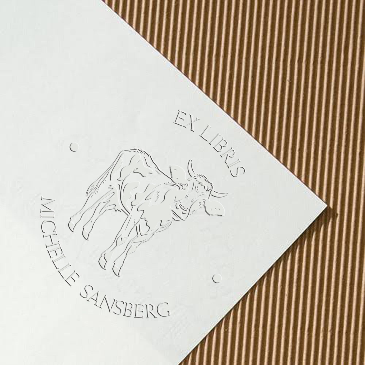 White embossed ex libris stamp seal featuring a bull, with silver-gray text, on a brown corrugated background.