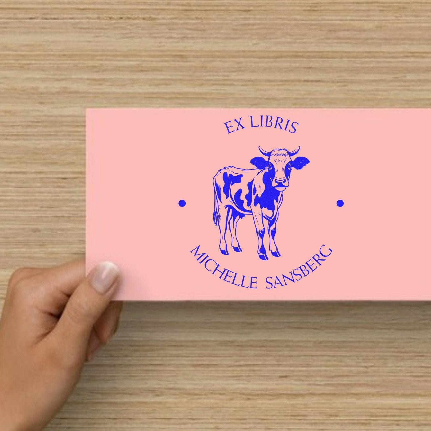Pink custom book stamp and seal with blue cow ex libris and Michelle Sansberg text, held by hand on a wood table.