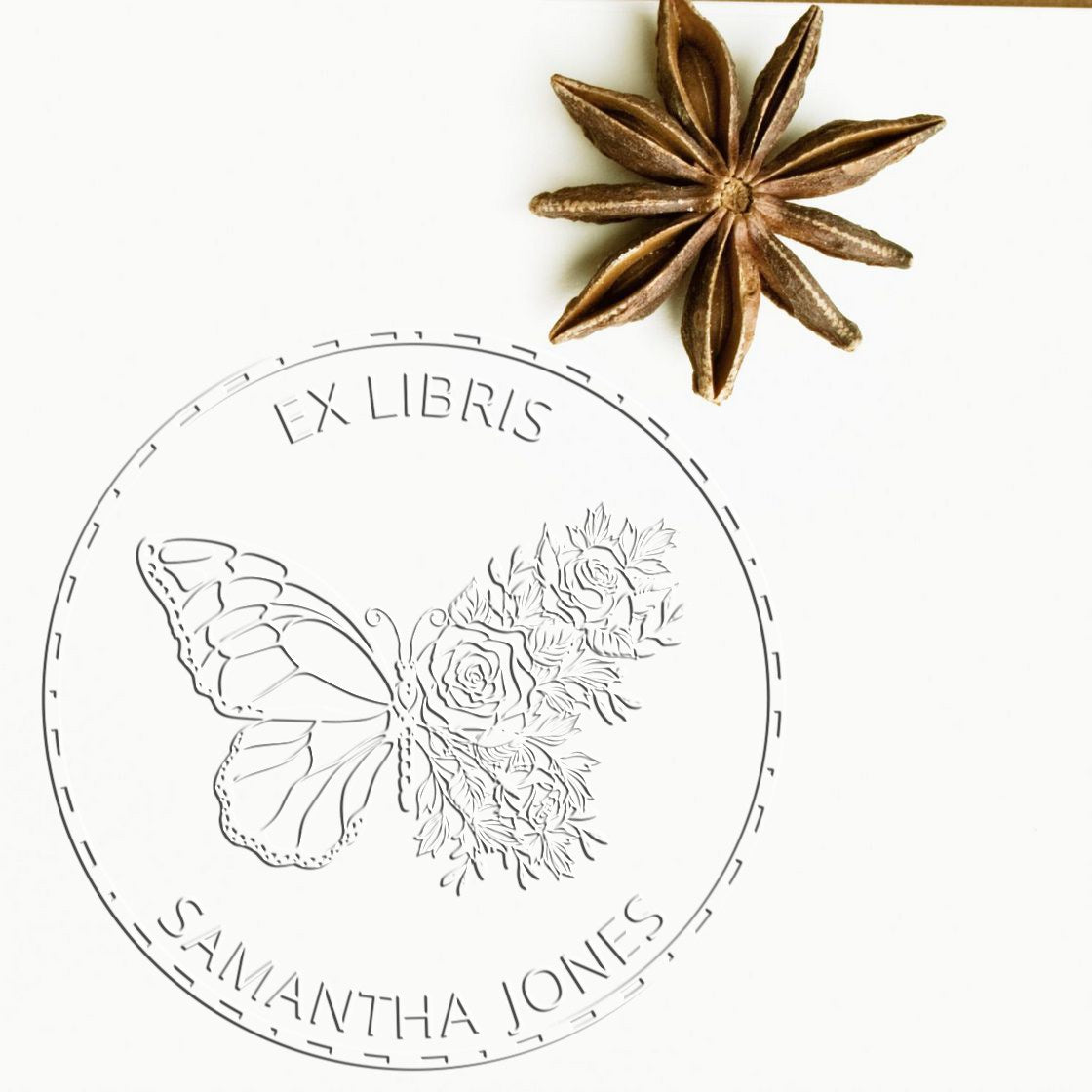 Silver embossing seal stamp with butterfly and roses, Ex Libris Samantha Jones, on white background with brown star anise.