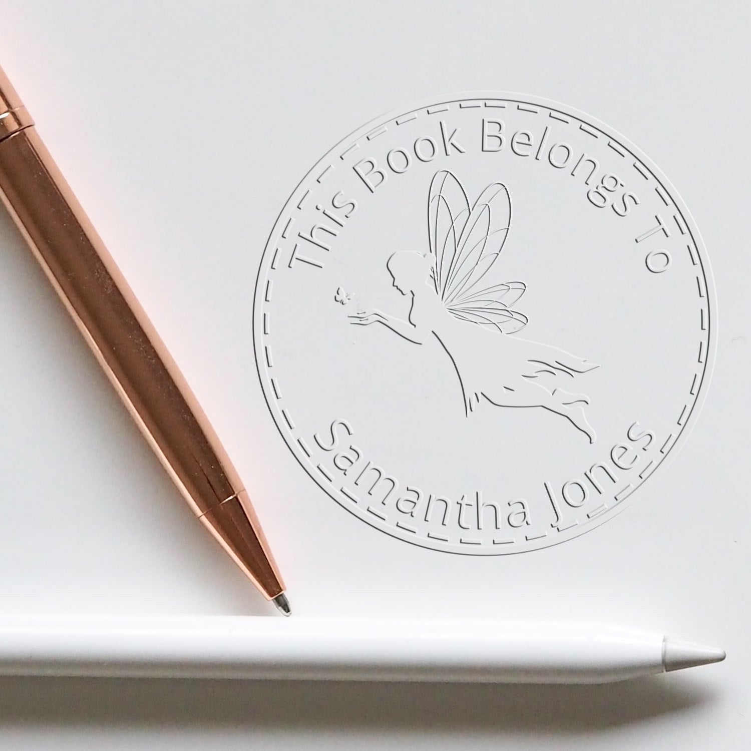 Custom white book ownership stamp with fairy seal, rose-gold pen, and white stylus; embossed text reads This Book Belongs To Samantha Jones.