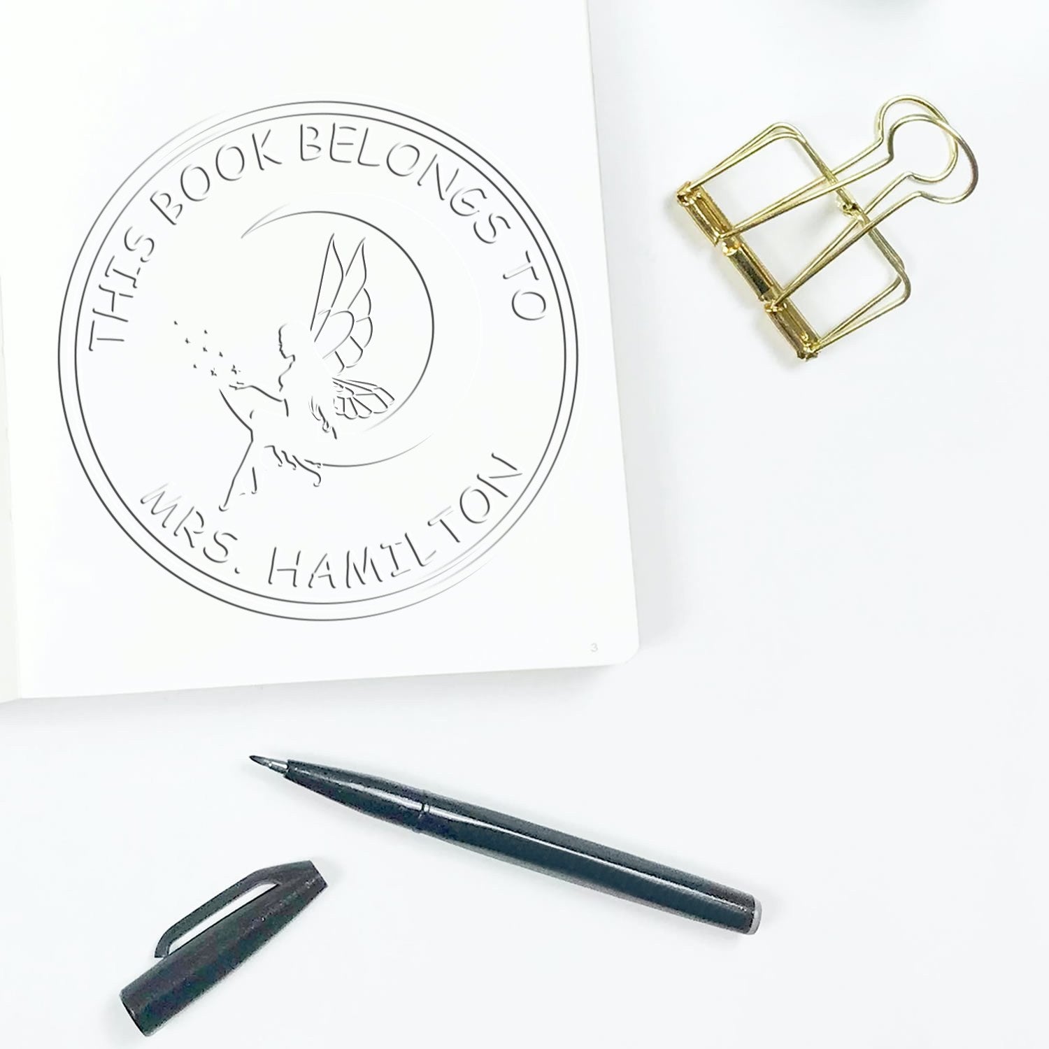Custom book stamp and seal with fairy design on white paper, plus gold binder clip and black pen on a clean white background.