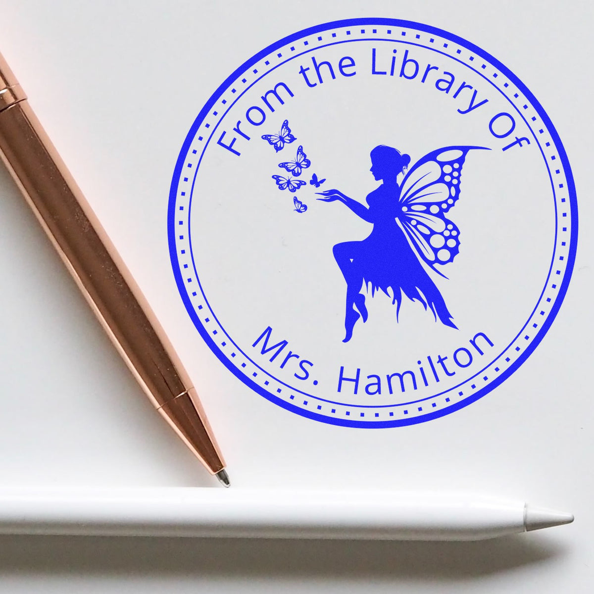 Blue circular library stamp seal with fairy and butterflies, reading From the Library Of Mrs. Hamilton, beside a rose-gold pen.