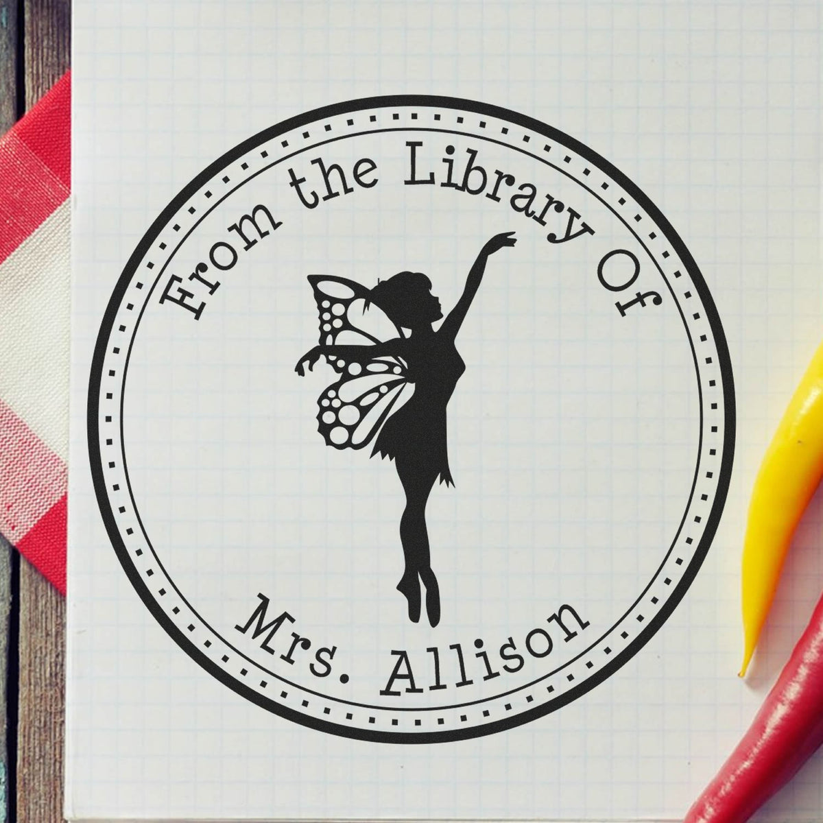 Black circular library stamp seal with fairy silhouette, From the Library Of Mrs. Allison, on pale grid paper with red and yellow pens.