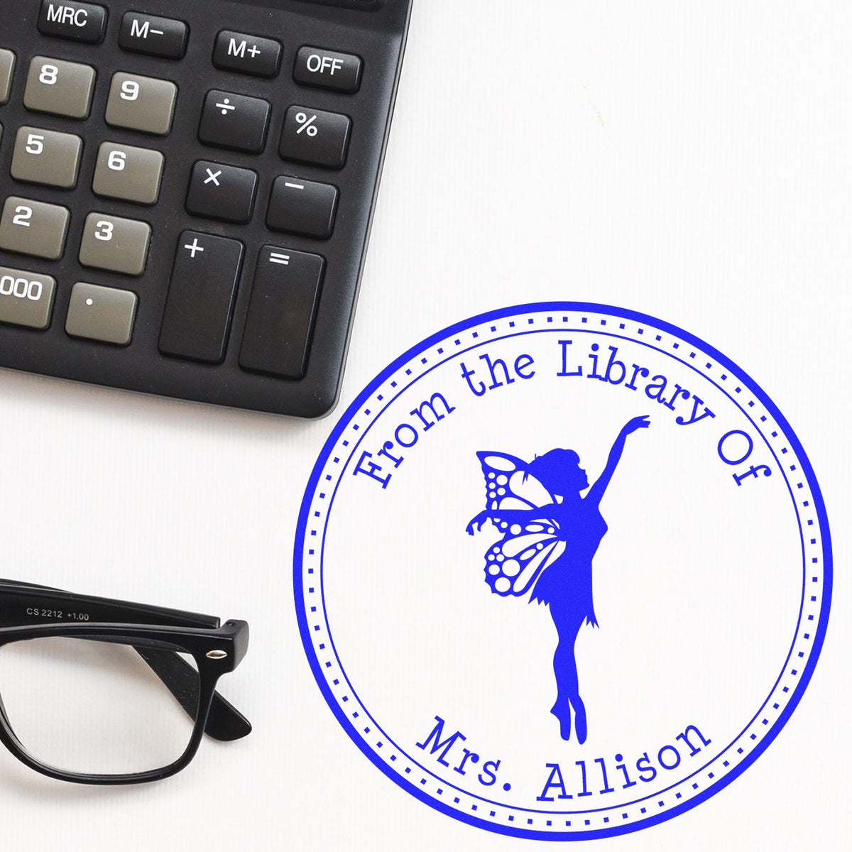 Blue custom library stamp seal with fairy silhouette and From the Library of Mrs. Allison, beside a calculator and glasses.