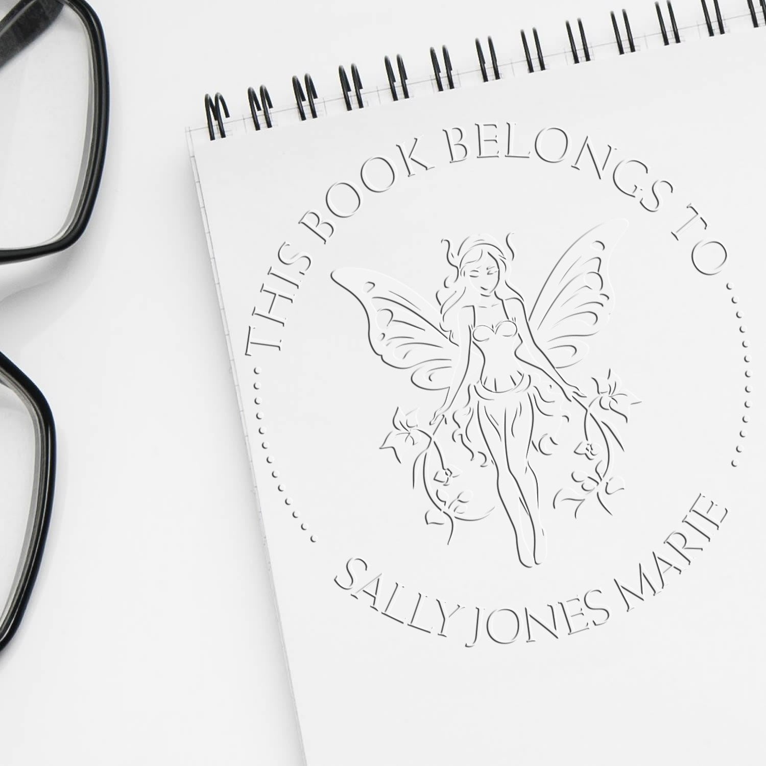 White custom book stamp and seal with fairy and embossed name on a spiral notebook, beside black eyeglasses.