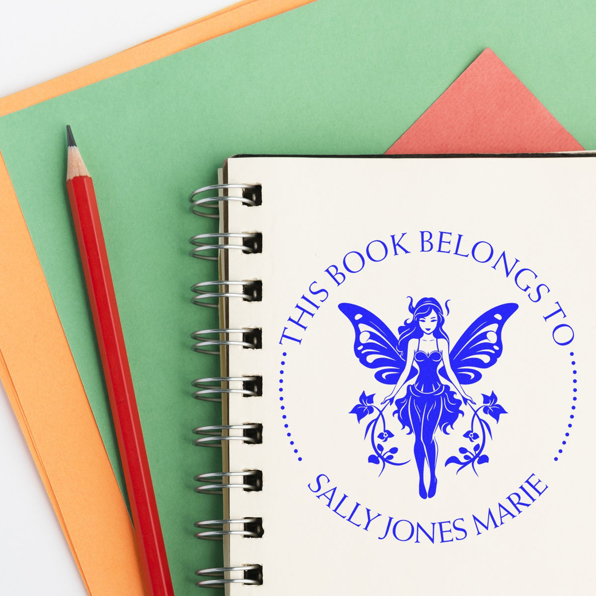 Blue fairy stamp seal on a spiral notebook beside a red pencil, with green, orange, and coral paper backgrounds.