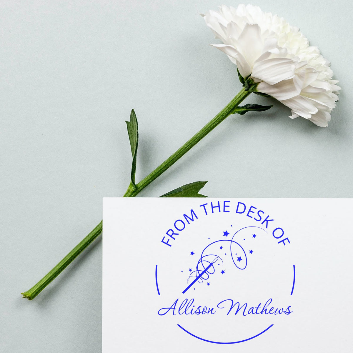 White flower beside a blue custom desk stamp and seal with Allison Mathews name on white paper.