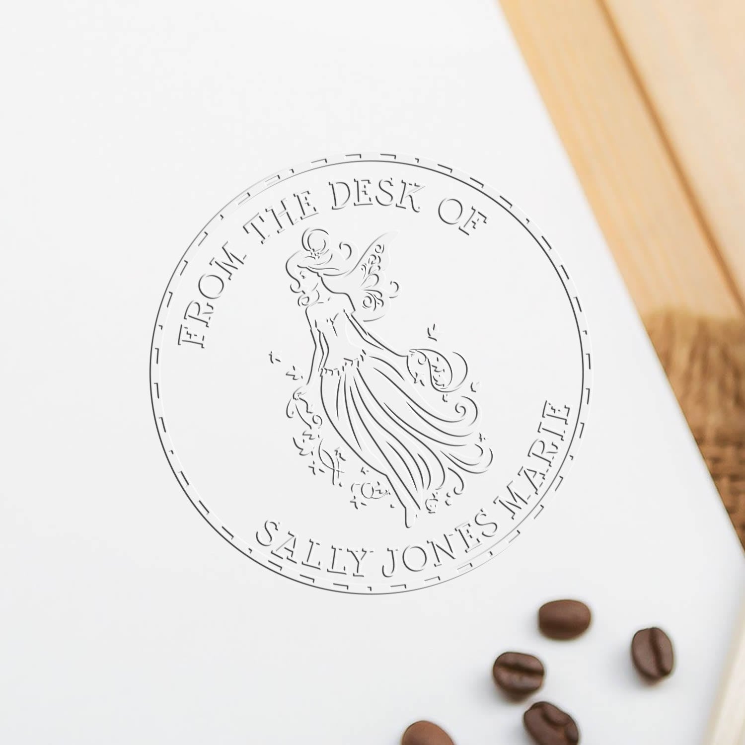 Embossed round desk stamp seal on white paper, featuring a fairy design, with coffee beans and warm wood accents.