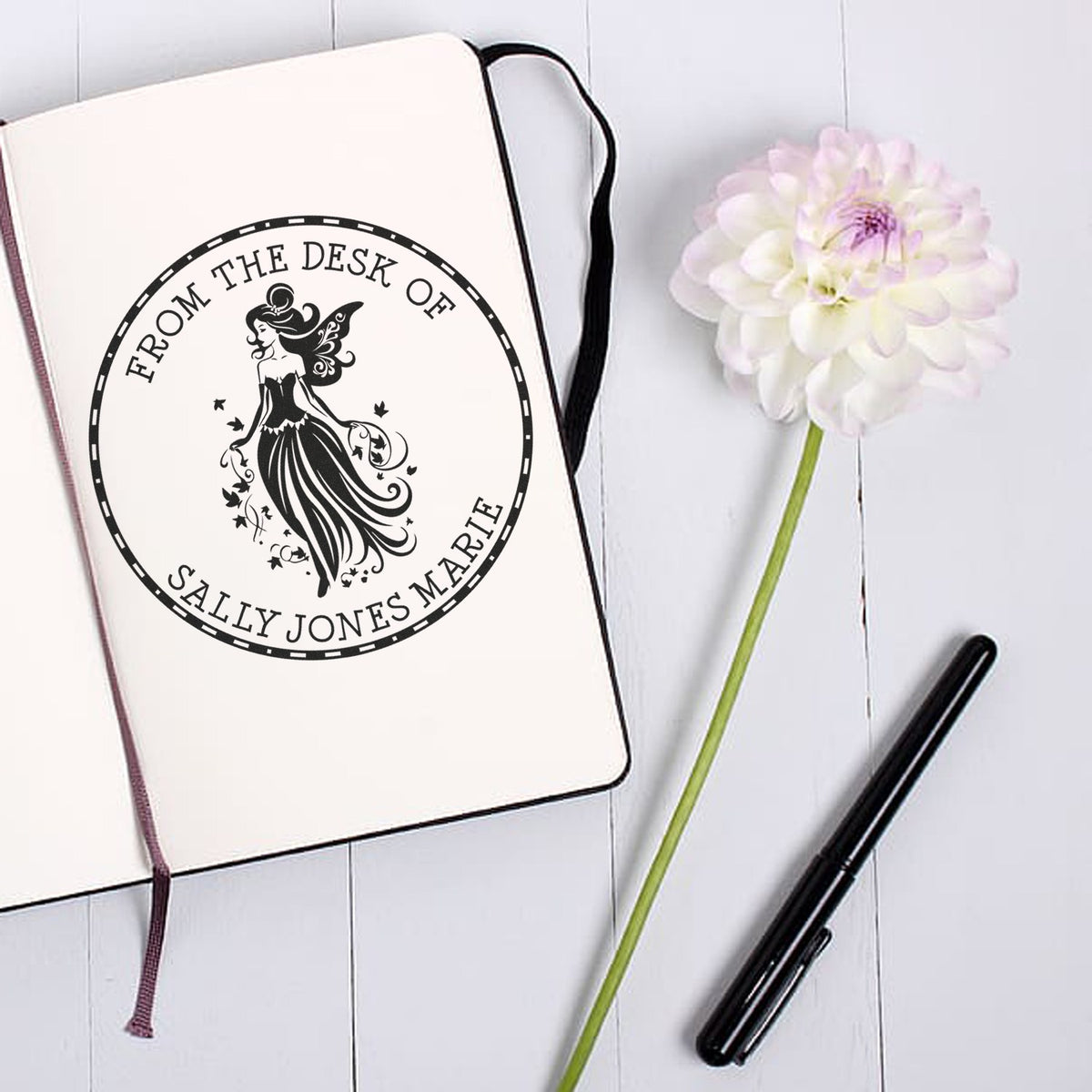 Custom black fairy stamp seal on cream notebook, reading From the Desk of Sally Jones Marie, beside a white flower and black pen.