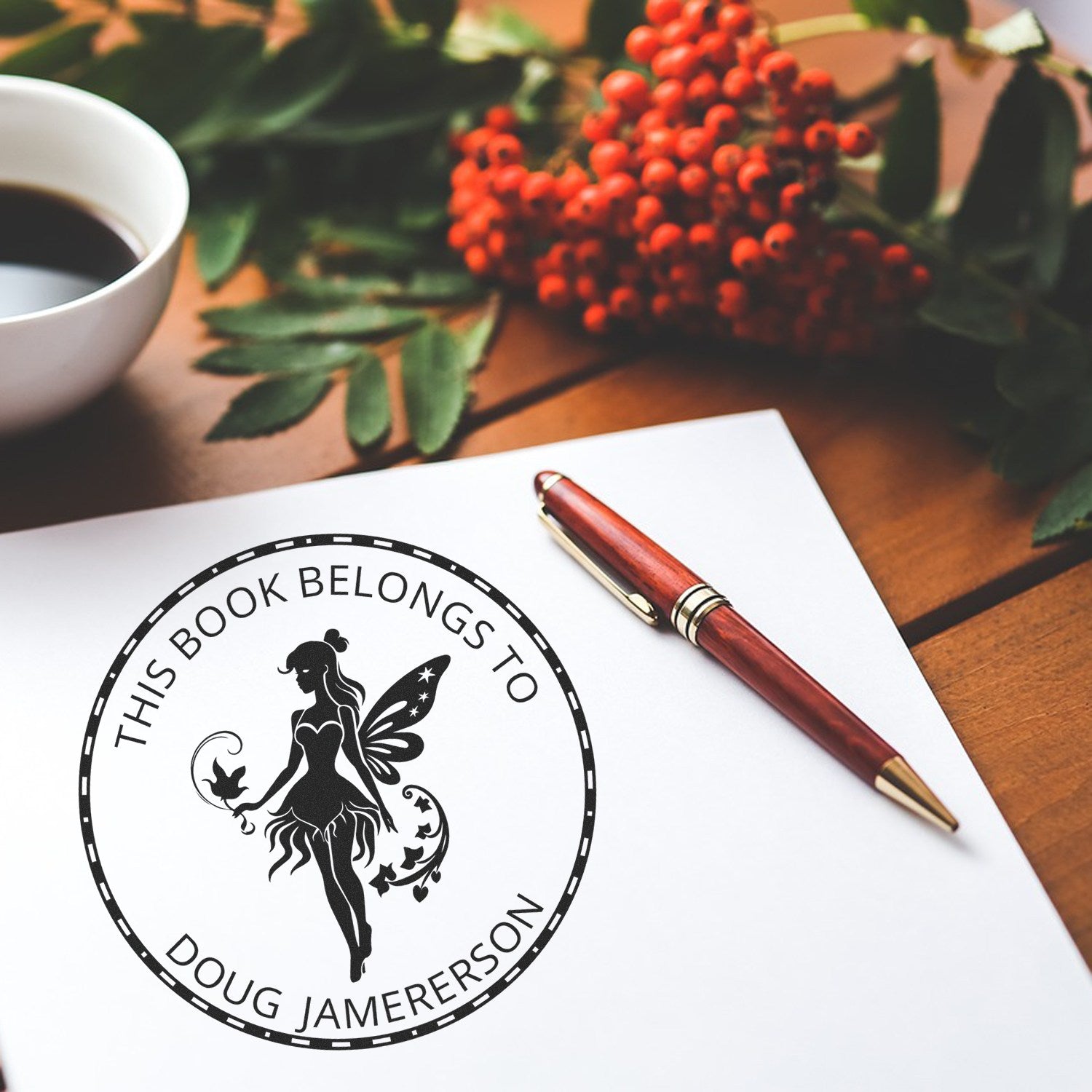 Custom book belongs to stamp seal with fairy silhouette on white paper, beside a burgundy pen, coffee cup, and red berries.