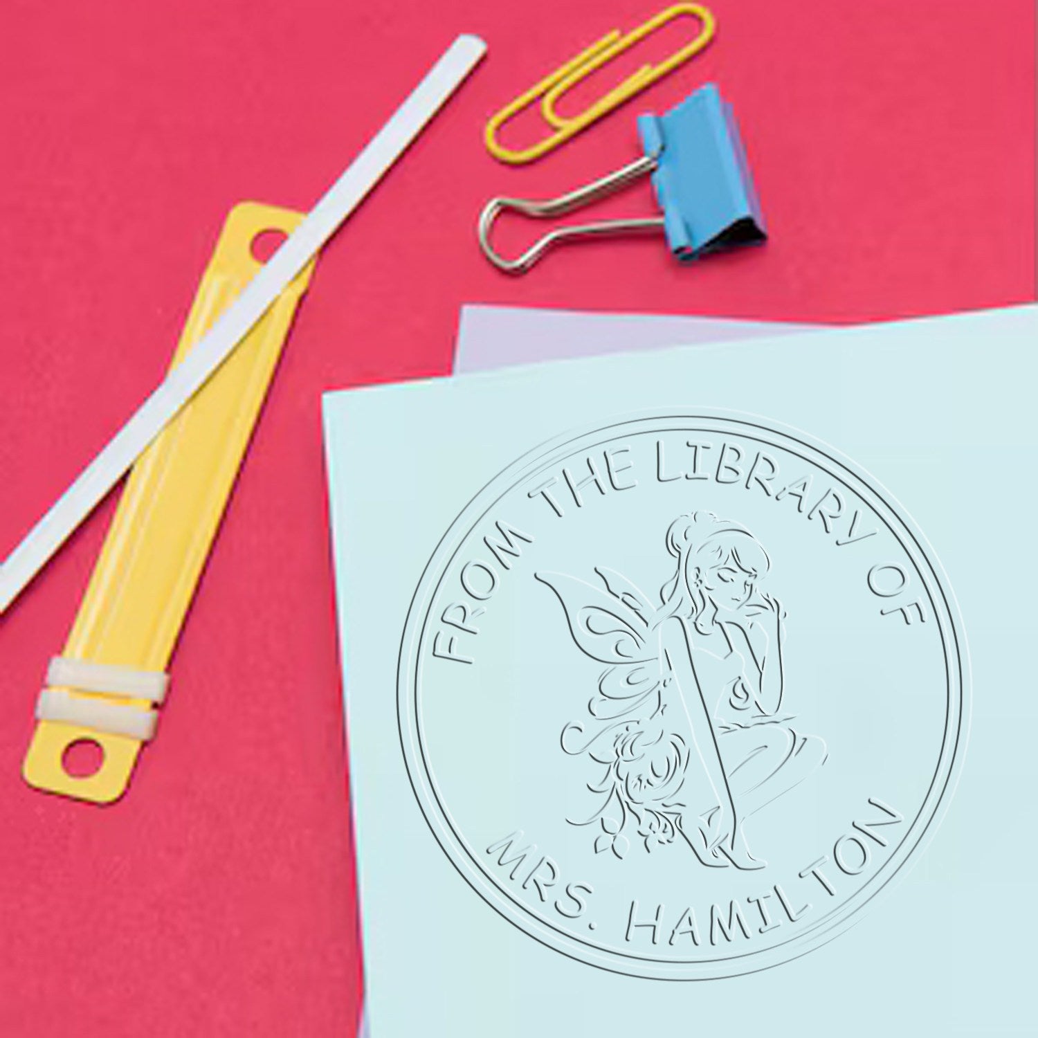 Embossed library seal stamp on pale blue paper, with colorful paper clips and binder clips on a bright pink background.