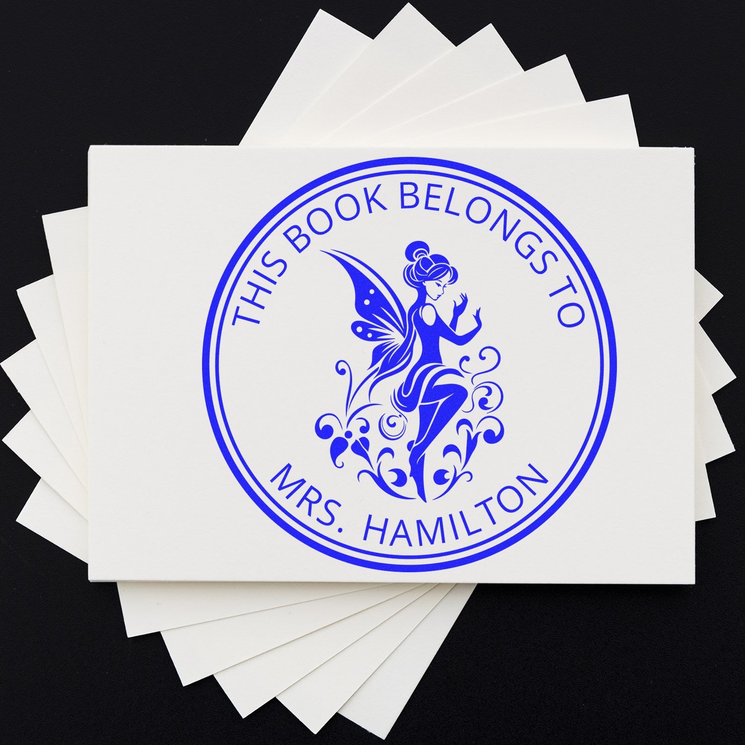 Blue custom bookplate stamp seal with fairy and circular This Book Belongs to Mrs. Hamilton design on white papers.
