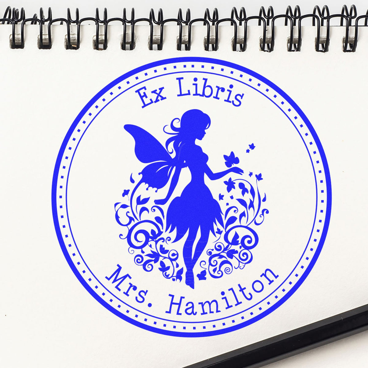 Blue Ex Libris stamp seal featuring a fairy silhouette and floral swirls on a white spiral-bound notebook page.