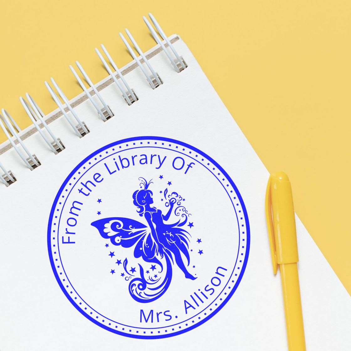 Blue circular fairy library return stamp seal on white spiral notebook, with a yellow pen on a yellow background.