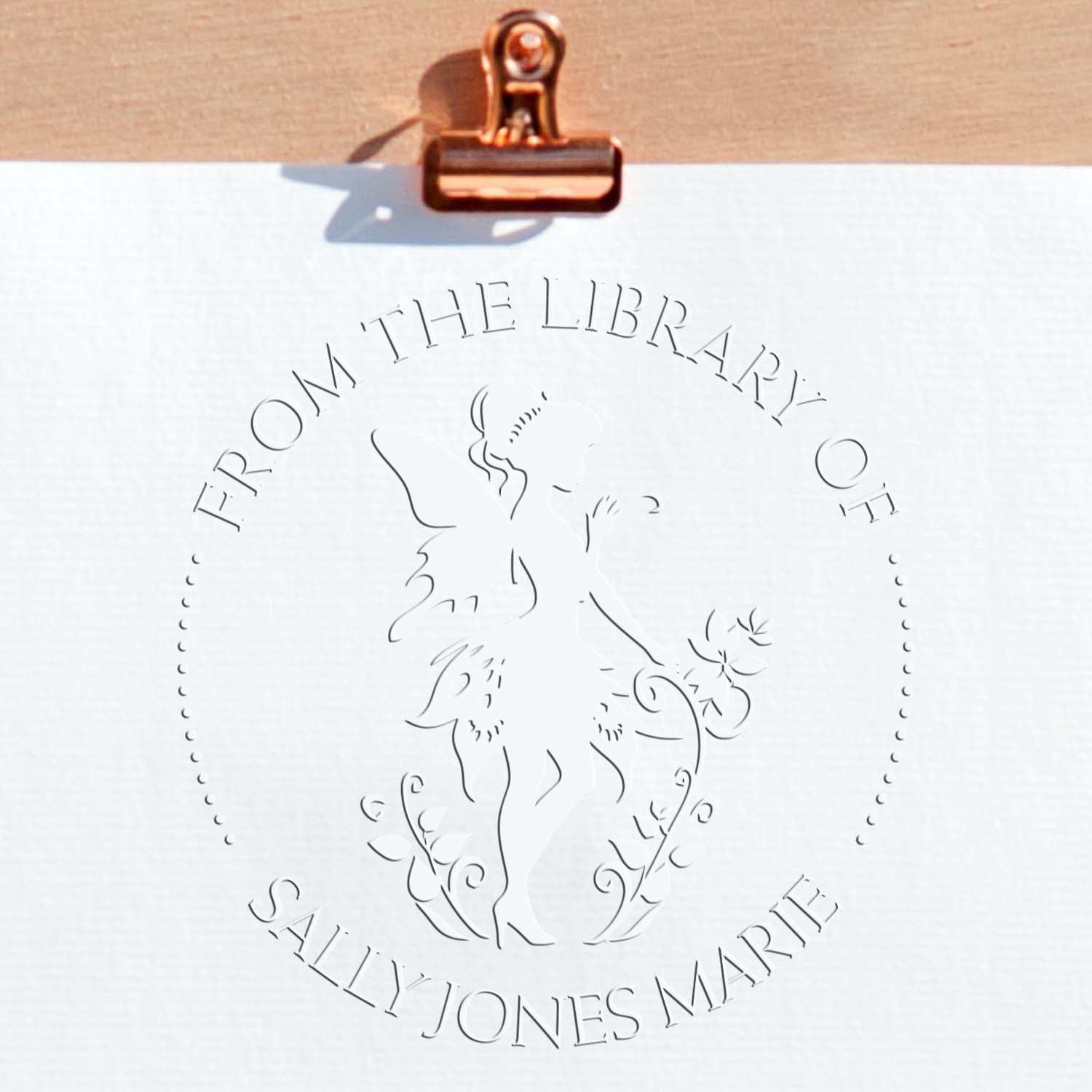 Copper binder clip holding white paper with embossed library seal reading From the Library of Sally Jones Marie and floral silhouette.