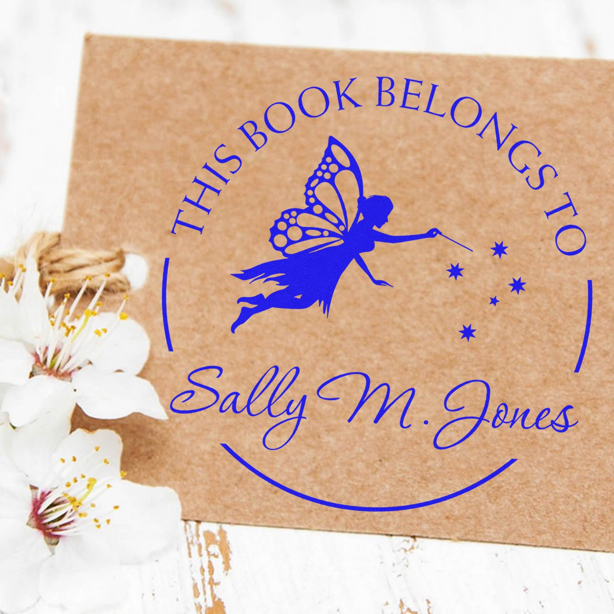Blue custom book seal stamp with fairy, stars, and Sally M. Jones on kraft paper, beside white blossoms.