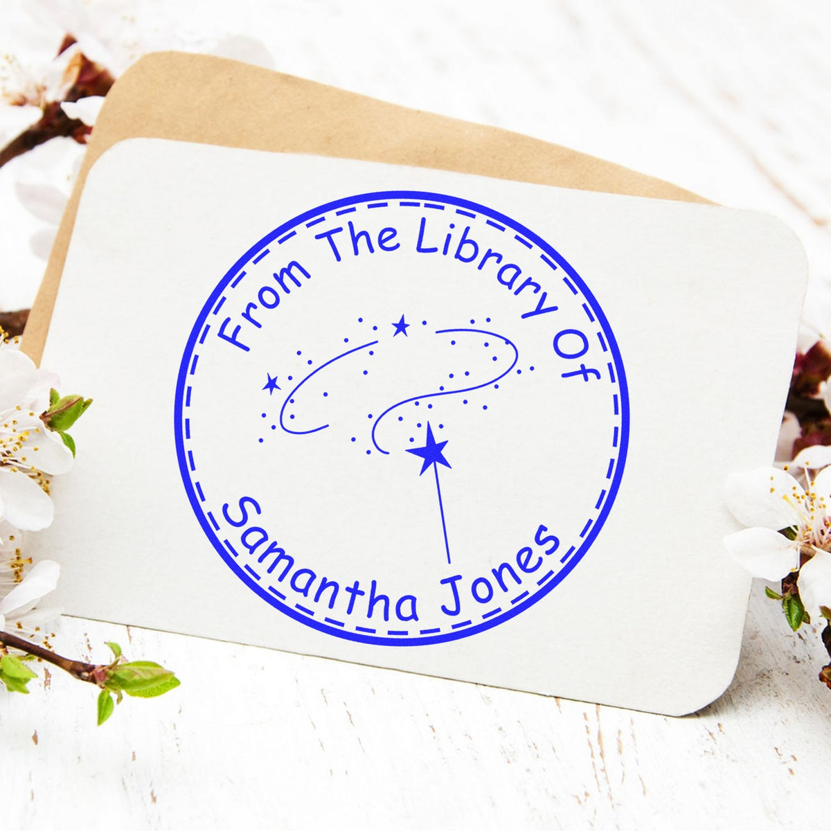 Blue circular From The Library Of Samantha Jones stamp seal on white card, with kraft envelope and blossoms.