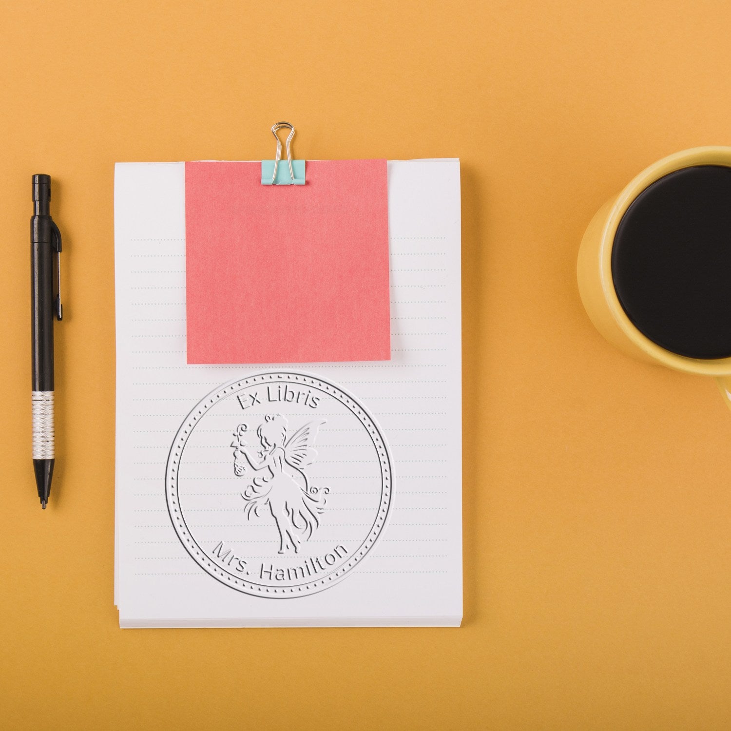 Custom ex libris stamp seal on lined notepad with pink clip, black pen, and coffee on mustard yellow background.