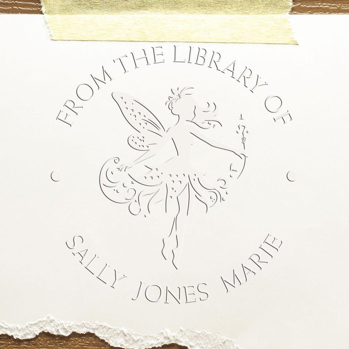 Embossed white library stamp seal with fairy silhouette and circular text on cream paper, held with masking tape.