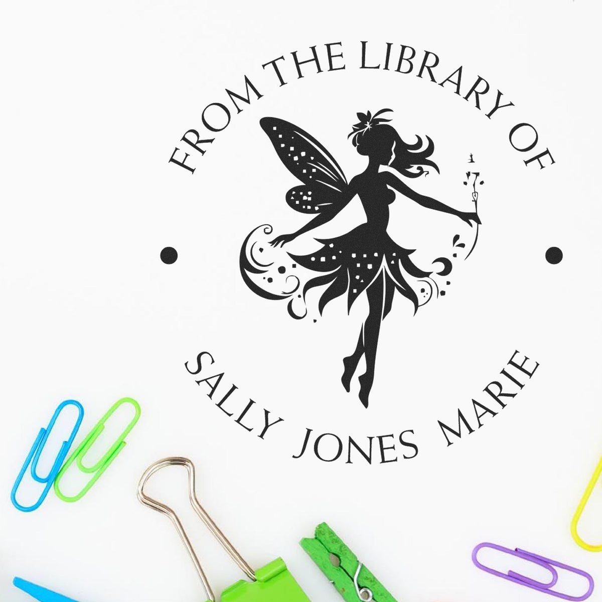 Black custom library stamp seal with fairy silhouette and circular text, on white background with colorful paper clips.