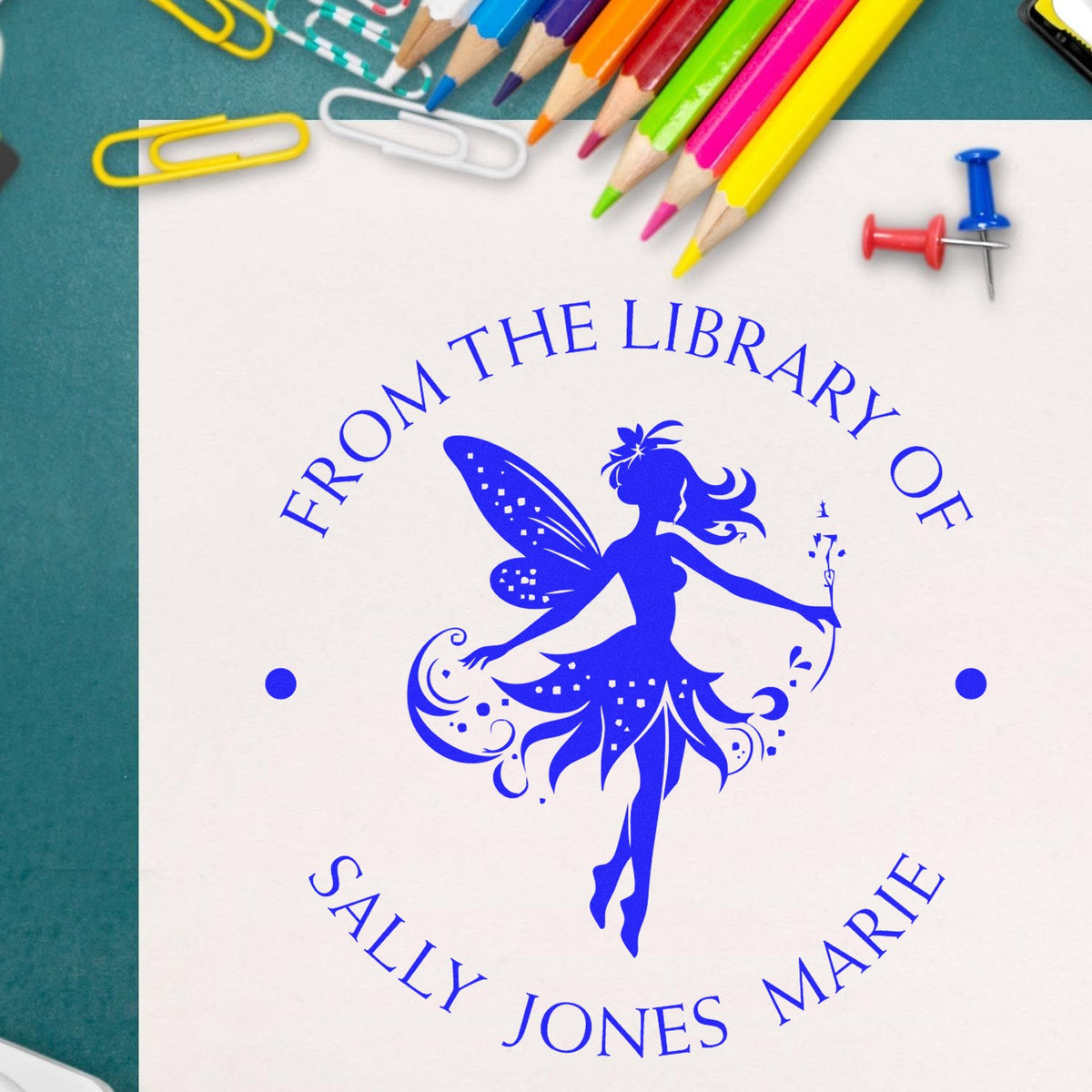 Blue custom library stamp seal with fairy silhouette on white paper, surrounded by colorful pencils, paper clips, and push pins.