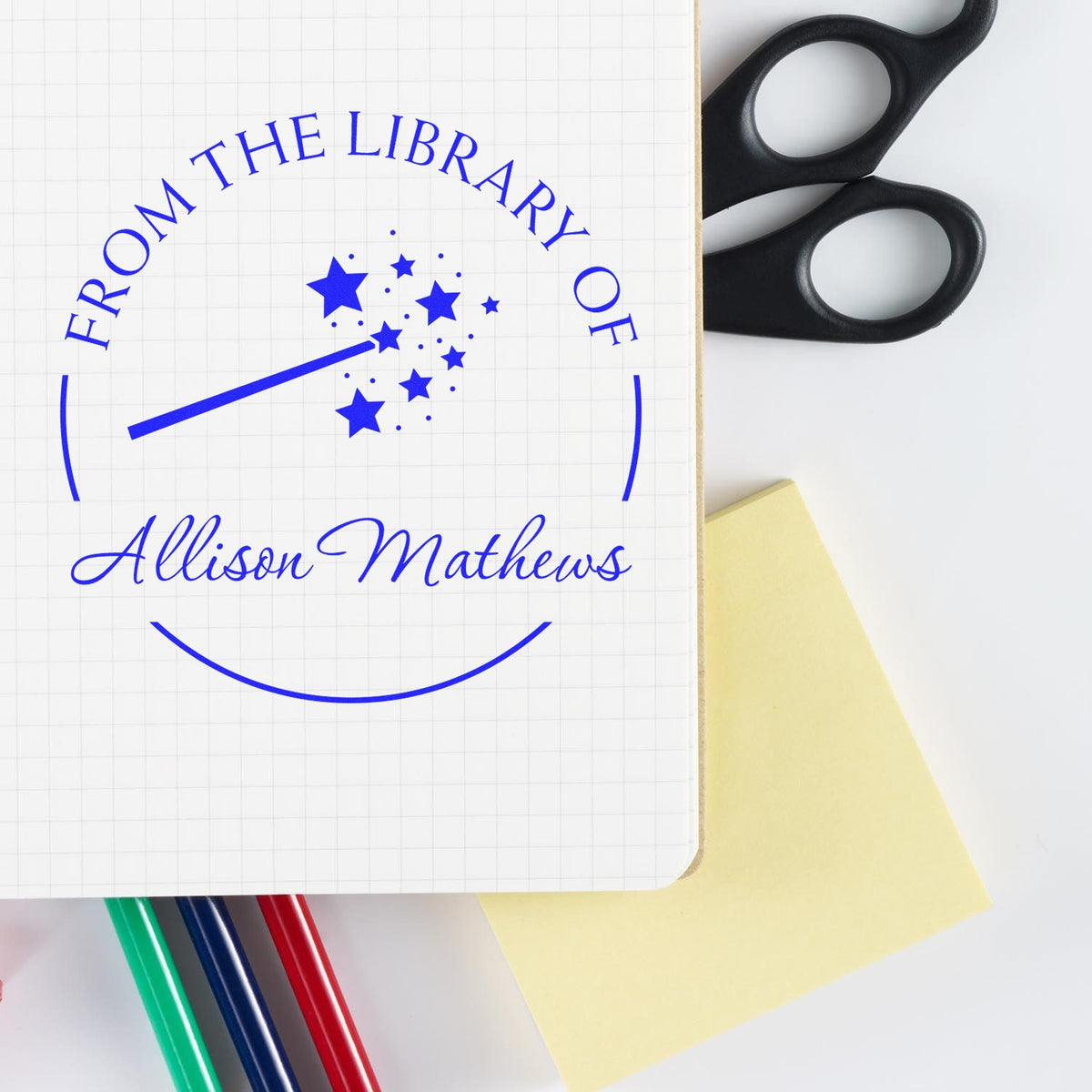 Blue custom library seal stamp on white grid paper, beside black scissors, yellow sticky note, and colored pens.