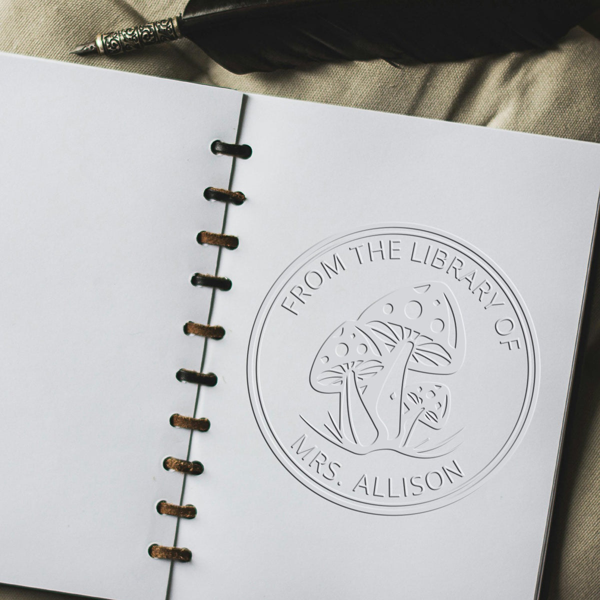 White spiral-bound booklet with embossed library seal stamp, mushroom logo, and black fountain pen.