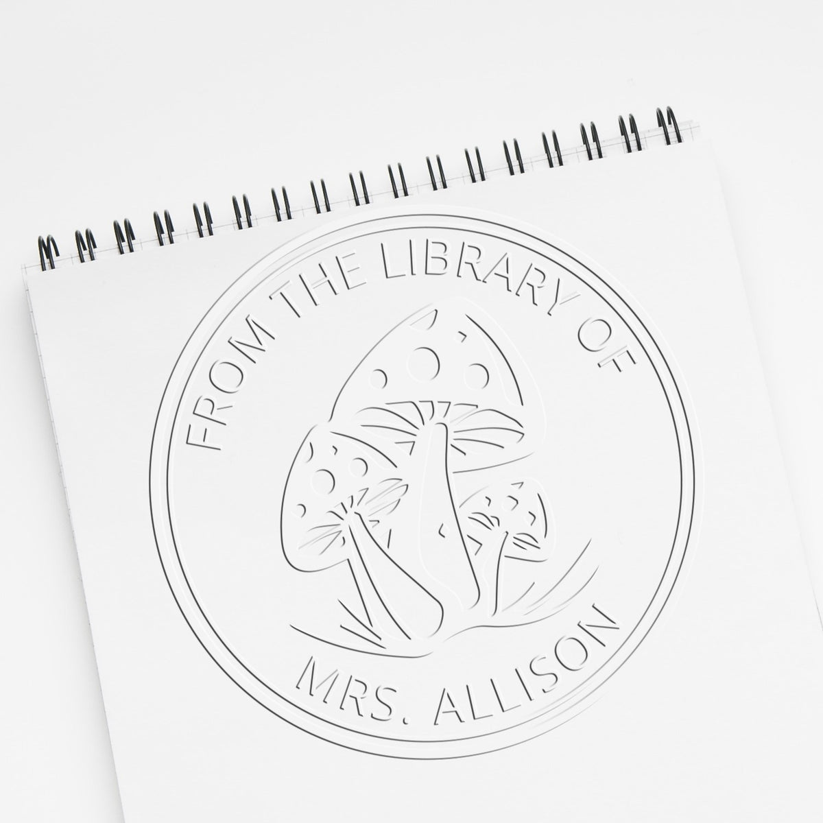 White embossed custom bookplate stamp seal on a spiral notebook, featuring mushrooms and text From the Library of Mrs. Allison