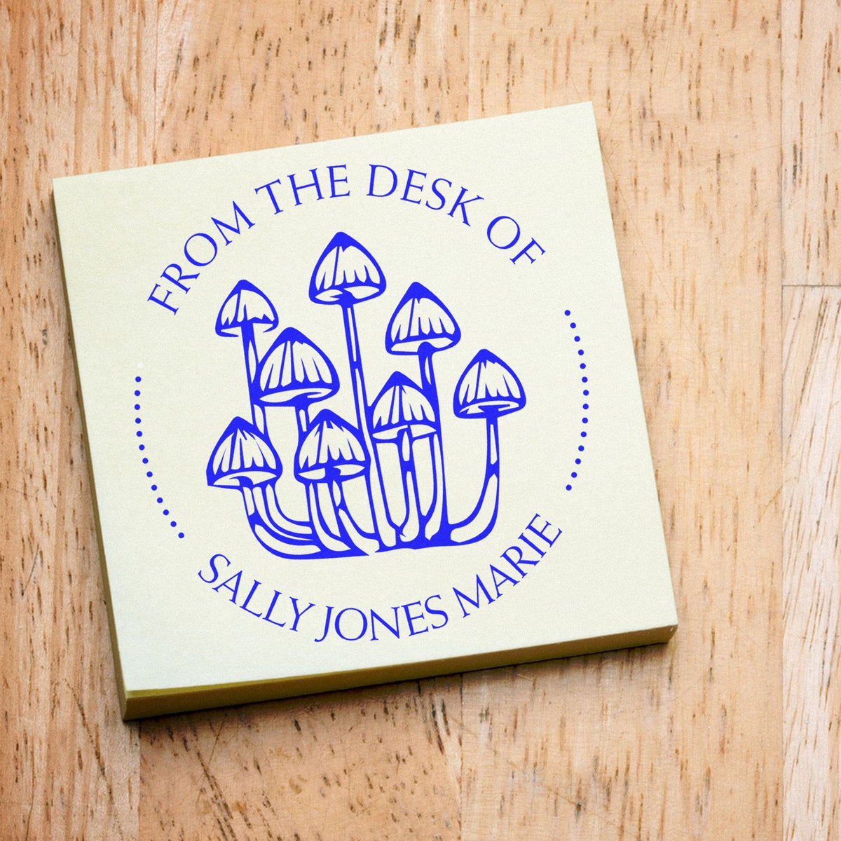 Personalized blue mushroom desk stamp seal on cream square, reading ĀFrom the Desk of Sally Jones Marie,Ā on wood background.