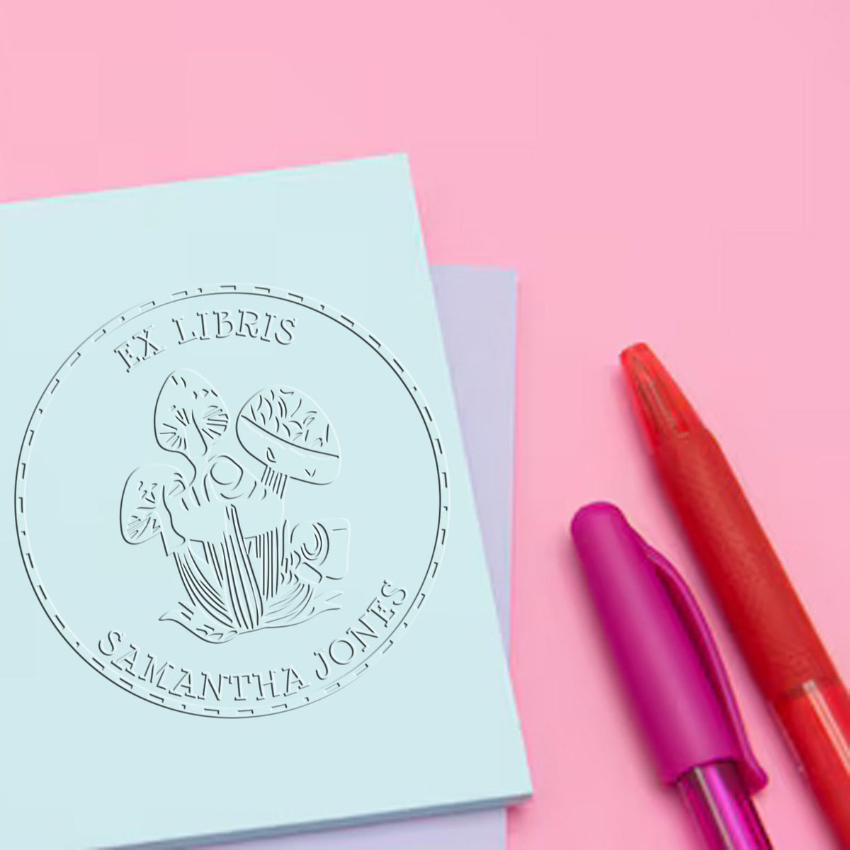 Custom stamp and embossing seal on pastel blue paper, with red and purple pens on a pink background.
