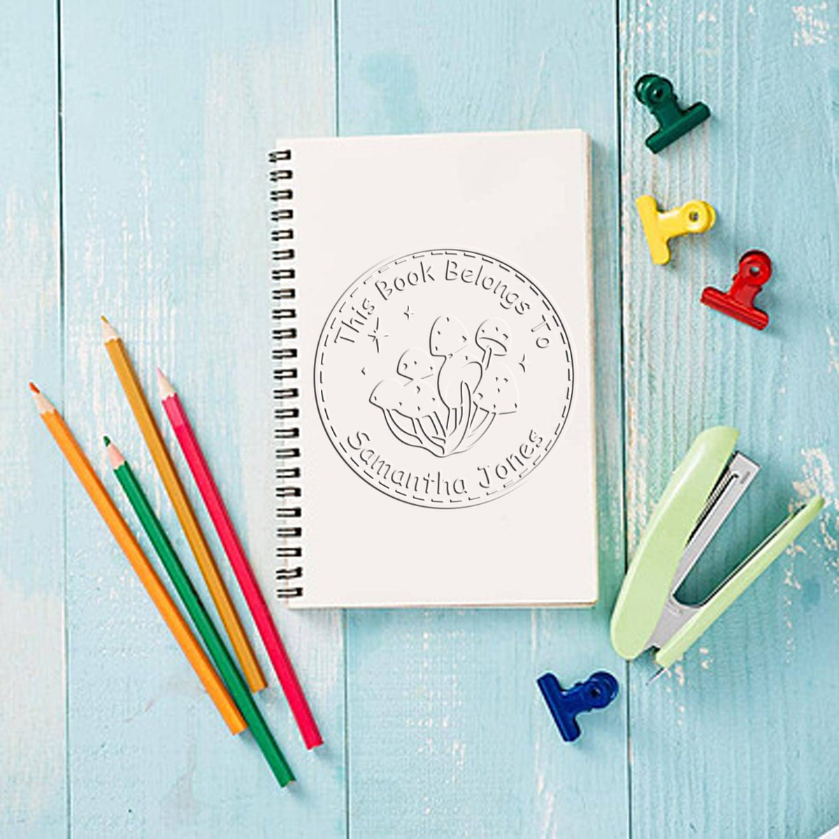 White spiral notebook with circular book-belongs-to stamp seal, colorful pencils, clips, and stapler on light blue wood.