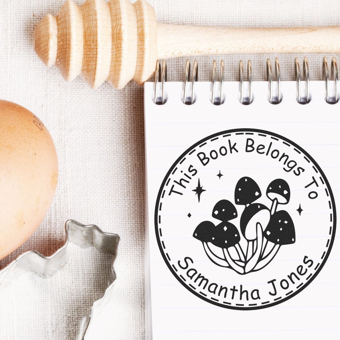 Black circular book stamp on notebook page with mushroom design and name Samantha Jones; wood-handled stamp and clear seal beside it.