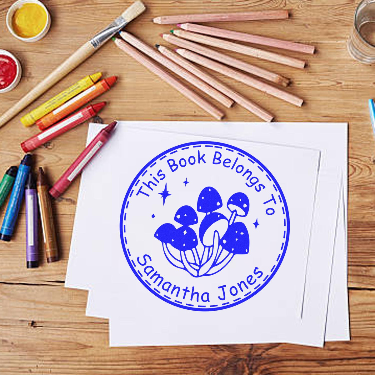 Blue custom library stamp and seal with mushroom design on white paper, surrounded by pencils on a wooden desk.