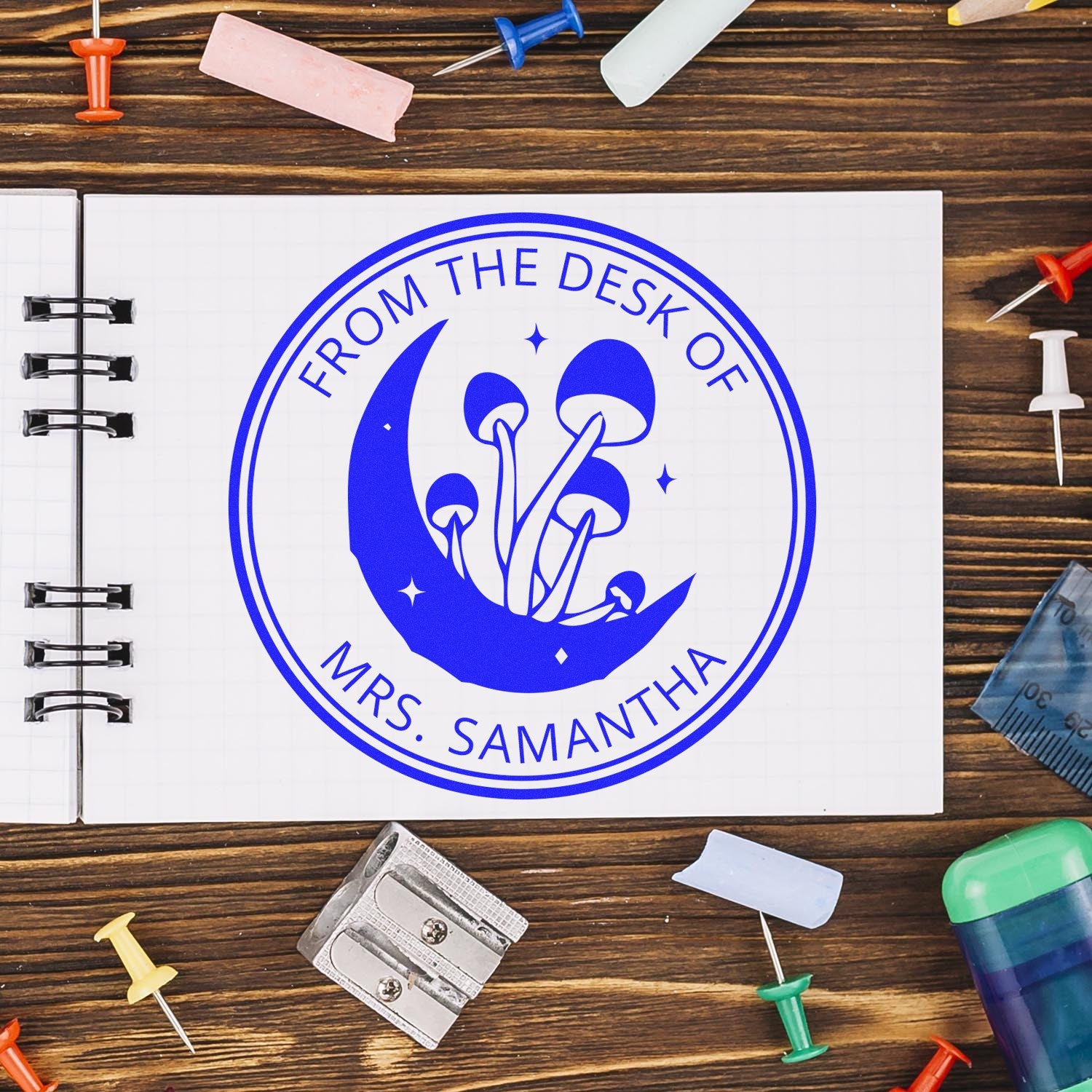 Blue custom stamp seal with mushrooms and crescent moon on a notebook, surrounded by colorful push pins and desk tools.