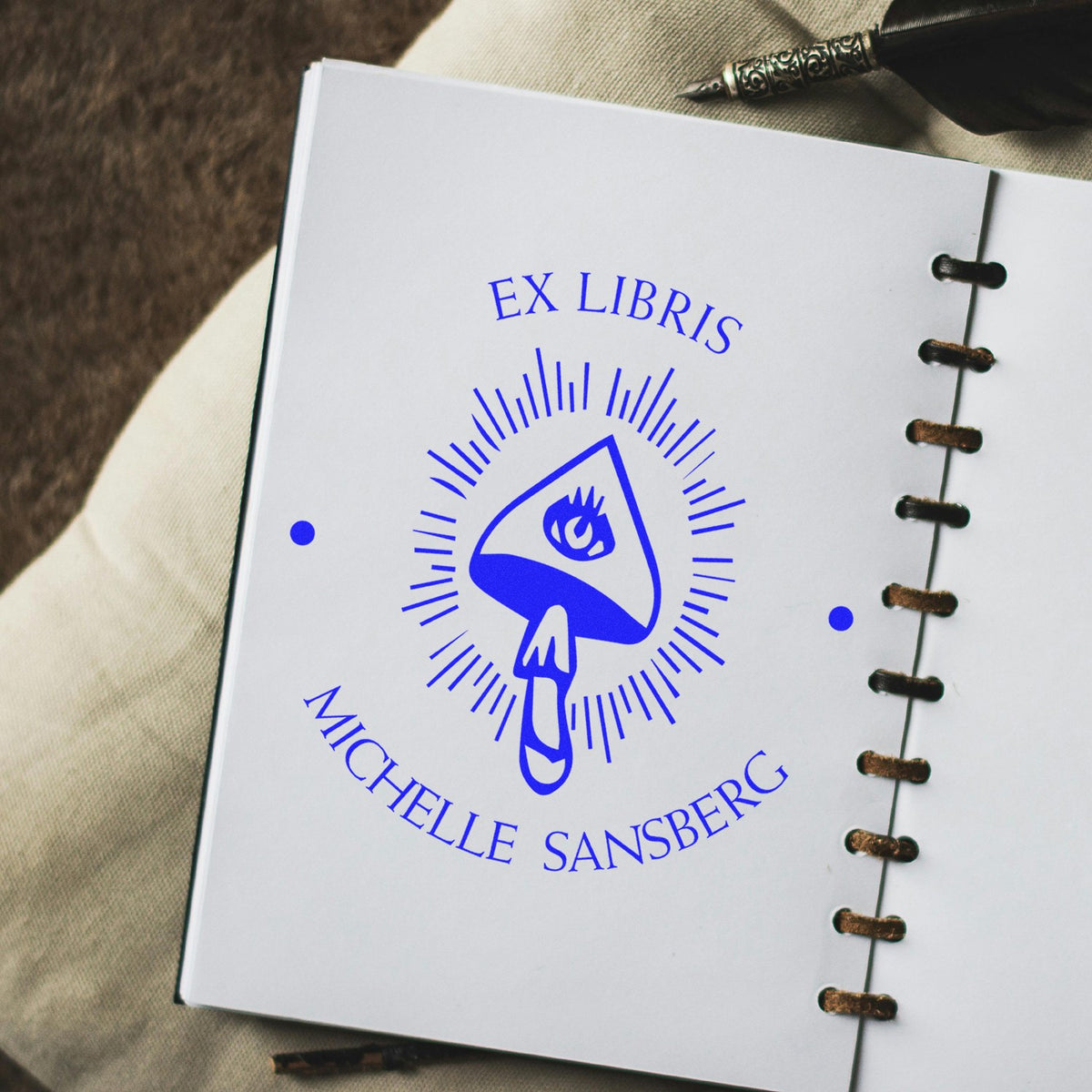 Blue ex libris mushroom stamp seal on a white notebook page with spiral binding and brown cord on beige fabric.